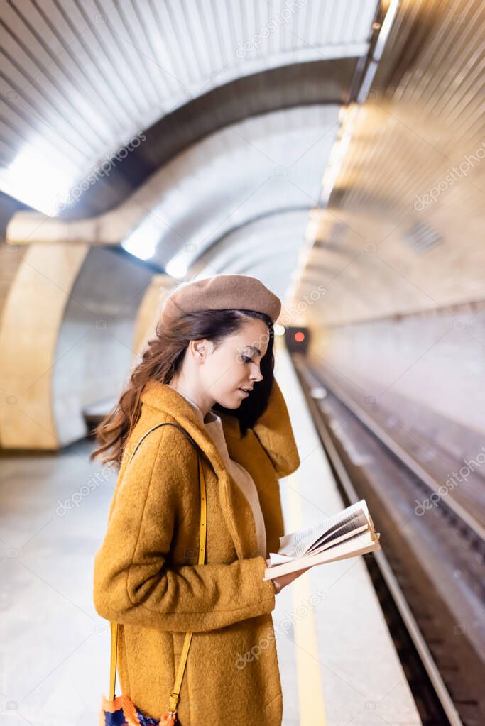young woman in coat and beret reading book on underground platform