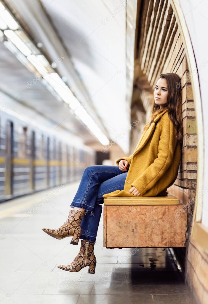 thoughtful woman in autumn outfit sitting on bench of metro platform with blurred train