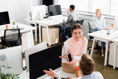 Akıllı telefon tutan neşeli iş kadını ofisteki bilgisayar ve meslektaşını işaret ediyor. 