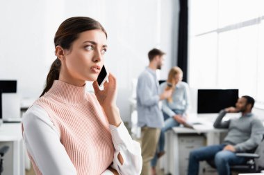 Genç iş kadını, ofisteki bulanık geçmişiyle akıllı telefondan iş arkadaşlarıyla konuşuyor. 