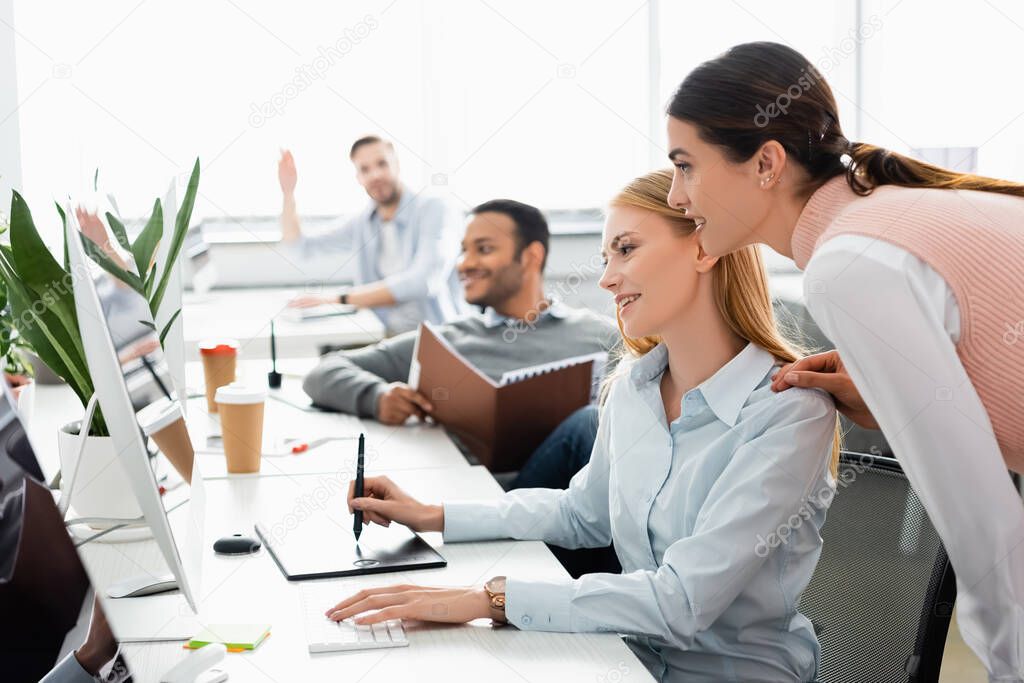 Smiling businesswoman using computer and graphics tablet near multiethnic colleagues on blurred foreground in office 