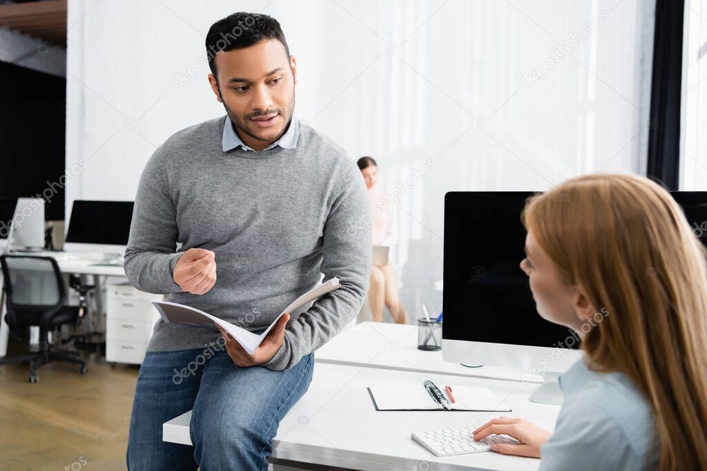 Indian businessman with paper folder talking to colleague on blurred foreground in office 