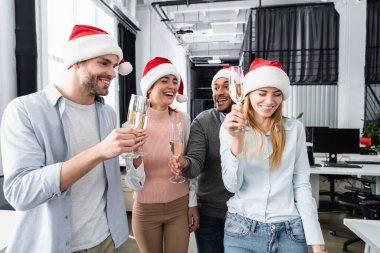 Ofiste ellerinde şampanya kadehleriyle Noel Baba şapkası takan pozitif etnik gruptan iş adamları. 