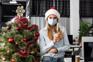 Sağlık maskeli ve Noel Baba şapkalı bir iş kadını. Ofiste yılbaşı ağacının yanında bir kadeh şampanya tutuyor. 