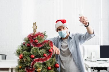 Sağlık maskeli bir iş adamı ve Noel Baba şapkalı elinde Noel ağacı yanında bir kadeh şampanya tutuyor. 