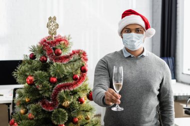 Sağlık maskeli Hintli bir işadamı ve Noel Baba şapkası ile Noel ağacının yanında bir bardak şampanya tutuyor. 