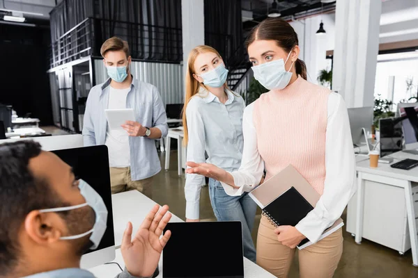 Businesswoman Medical Mask Notebooks Pointing Indian Colleague Blurred Background Office — Stock Photo, Image