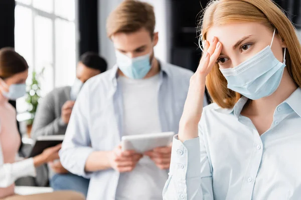 病気ビジネス女性で医療マスクタッチ頭近く同僚上のぼやけた背景上のオフィス — ストック写真
