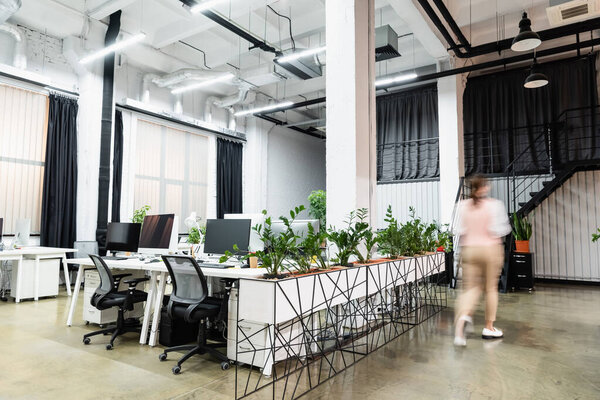 Motion blur of businesswoman walking near computers and plants in office 