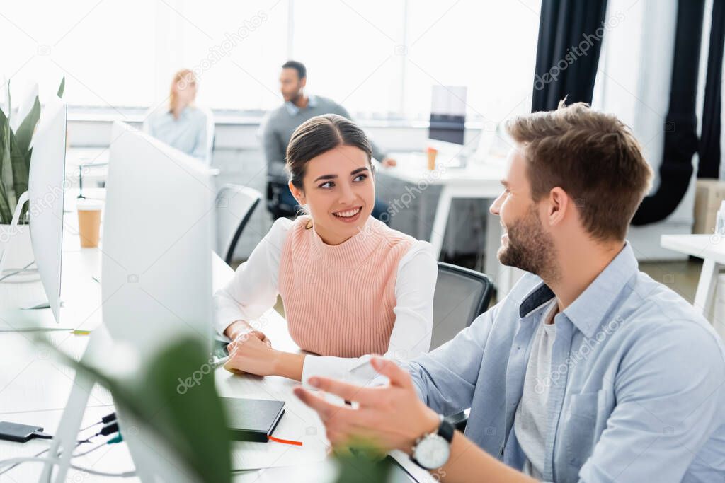 Smiling businesspeople talking near computers and notebook on table in office 