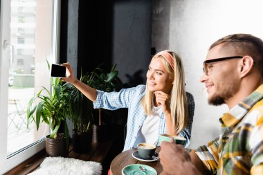 Süslü sarışın kadın kahve evinde arkadaşıyla cep telefonundan selfie çekiyor.