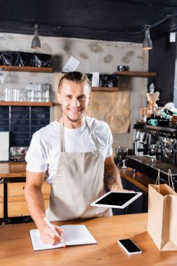 Neşeli, yakışıklı barista, not defterine yazarken kameraya bakıyor ve boş ekran ile dijital tableti tutuyor. 