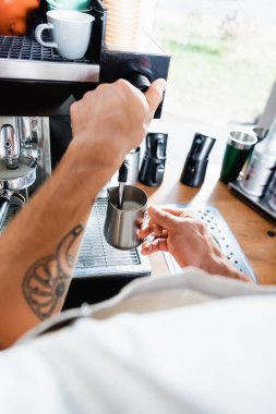 Ön planda kahve makinesinin buharlı buharlı buharlı buharlı sütlü metal bardak tutan dövmeli barista görüntüsü.