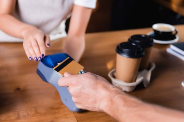 Adamın kredi kartıyla ödeme yaptığını gösteren kısmi bir bakış açısı kahvenin yanında ve bulanık arka planda barista var.
