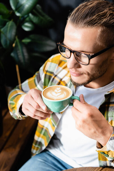 high angle view of young man in eyeglasses holding cup of coffee with latte art