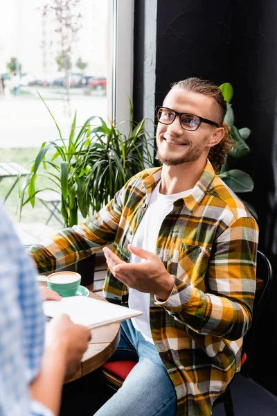 Gözlüklü Gülümseyen Genç Adam Bulanık Önplanda Garsonun Yanında Parmağıyla Işaret — Stok fotoğraf