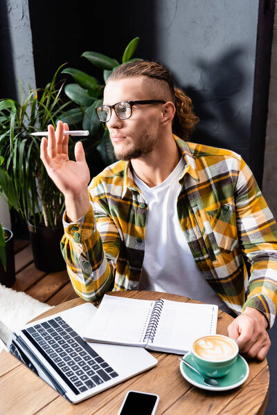вдумчивый фрилансер в клетчатой рубашке с ручкой рядом с ноутбуком, ноутбуком и чашкой кофе с искусством латте