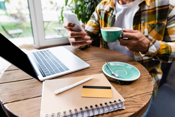 cropped view of freelancer chatting on smartphone and holding cup of coffee near laptop, notebook and credit card in cafe, blurred background