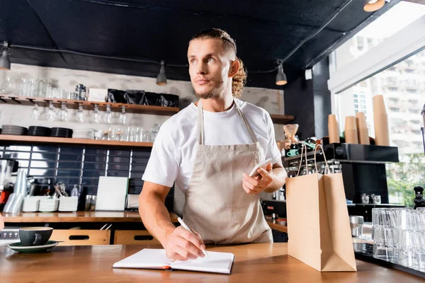 Young Barista Holding Smartphone While Writing Notebook Paper Bag Bar — Stock Photo, Image