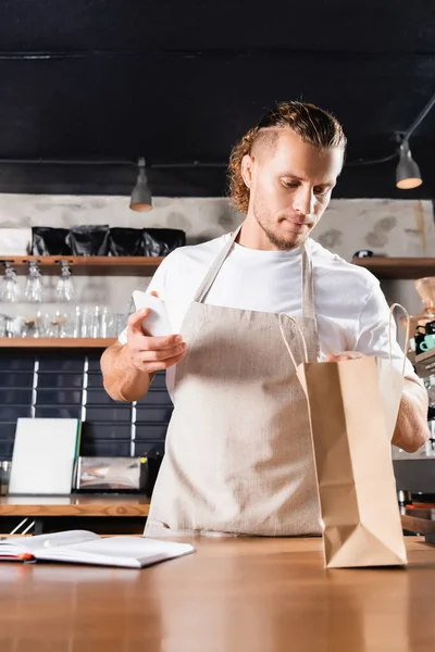 Handsome Barista Apron Looking Paper Bag While Holding Smartphone Notebook — Stock Photo, Image