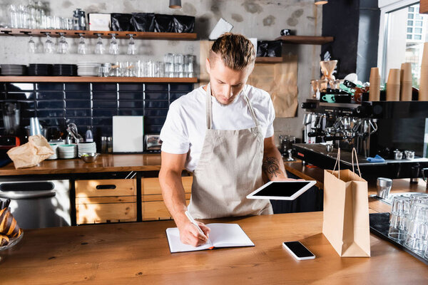 young waiter writing in notebook while holding digital tablet near smartphone and paper bag