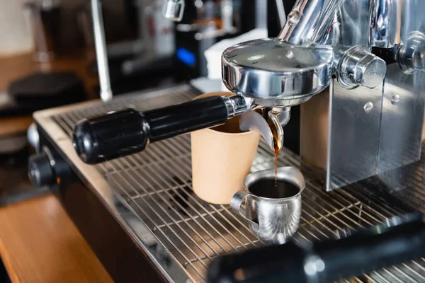 metallic mug with coffee near dispenser of professional coffee machine and disposable cup, blurred foreground