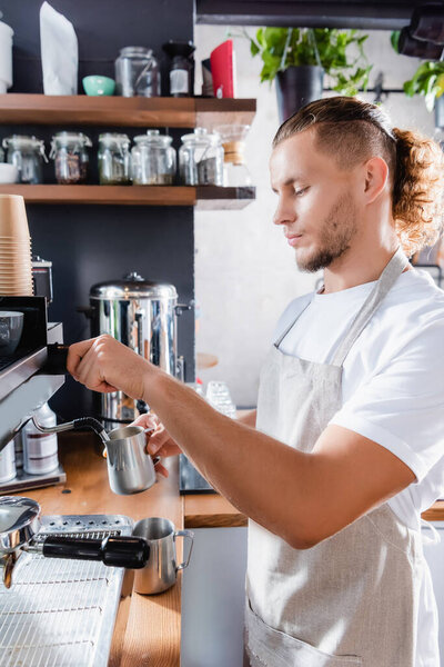 young barista in apron holding metallic milk mug near coffee machine steamer