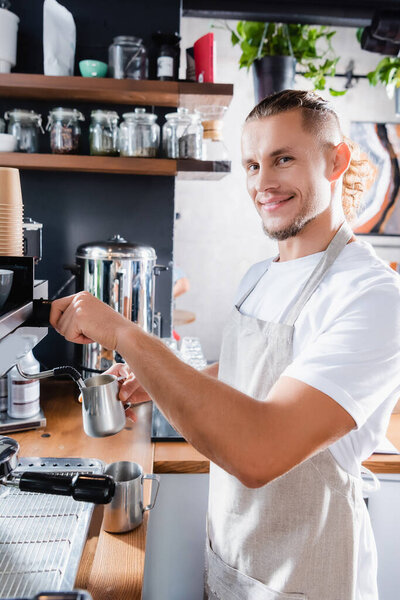 young smiling barista looking at camera while holding metallic milk mug near steamer of coffee machine