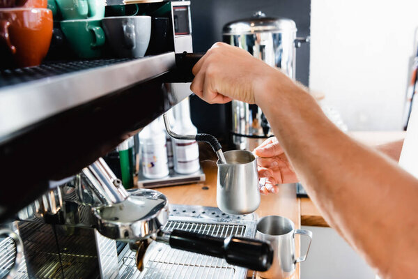 cropped view of barista holding metallic milk mug near steamer of coffee machine