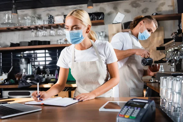 Jovem Garçom Máscara Médica Escrevendo Notebook Perto Laptop Barista Trabalhando — Fotografia de Stock