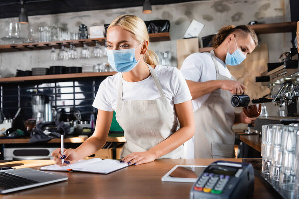 young waiter in medical mask writing in notebook near laptop, and barista working on background