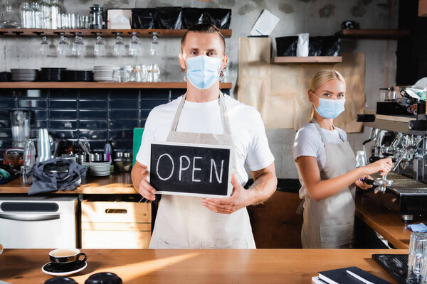 waiter in medical mask holding board with open lettering near barista working on background