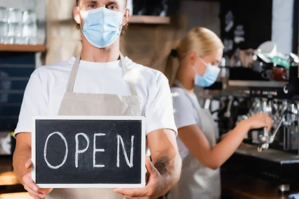 waiter in medical mask holding board with open lettering near barista working on blurred background