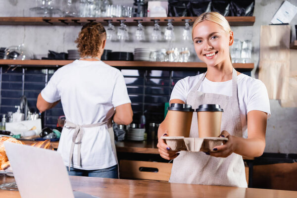 cheerful blonde barista holding coffee to go near colleague on background