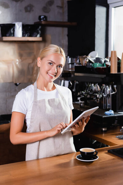 smiling blonde barista using digital tablet near cup of coffee on bar counter