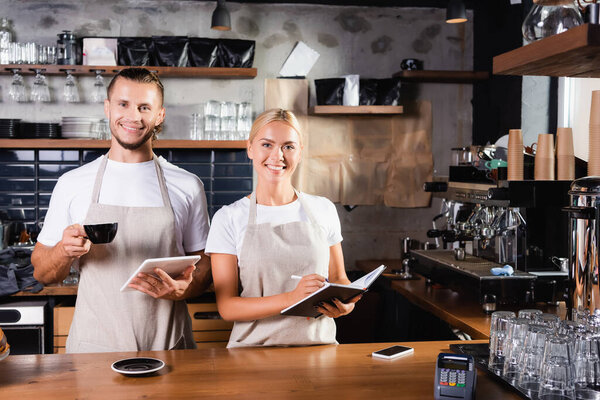 young baristas in aprons smiling at camera while standing at bar counter