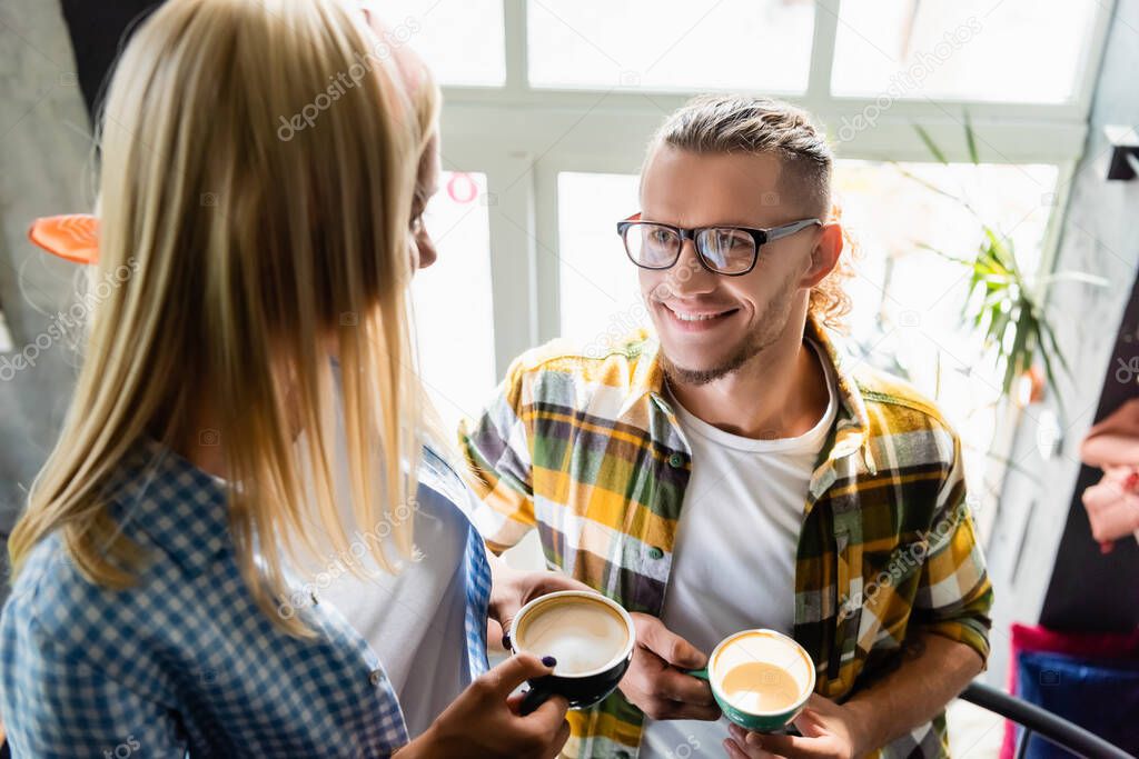 smiling young man in eyeglasses and woman on blurred foreground holding cups of coffee in cafe