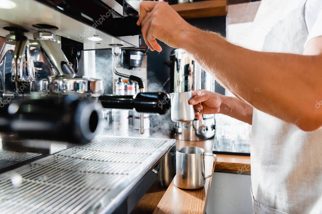 partial view of barista holding metallic milk mug near steamer of coffeemaker on blurred foreground