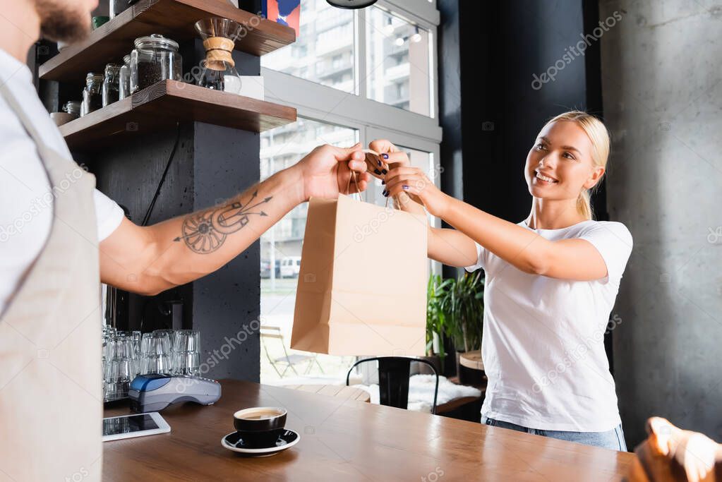 smiling blonde woman taking paper bag from tattooed barista on blurred foreground