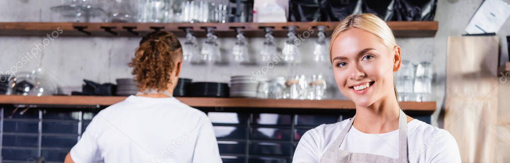 Young blonde barista smiling at camera near colleague on background, banner — Stock Image Young Blonde Barista Smiling Camera Colleague Background Banner Stock Image