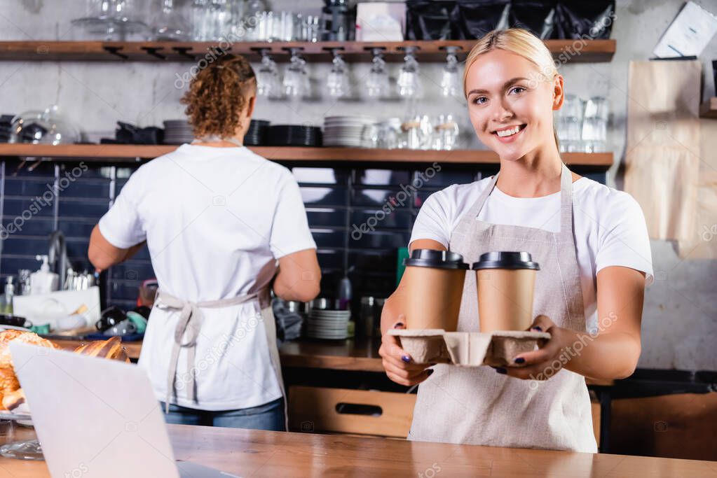 cheerful blonde barista holding coffee to go near colleague on background