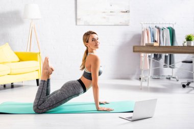 Evde laptopun yanında yoga yapan gülümseyen sporcu kadının yan görüntüsü bulanık ön planda. 