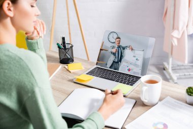 İşadamı, Webinar 'ın bulanık ön planı sırasında deftere yazan serbest bilgisayarın yanındaki ekranda parmak ucu ile işaret ediyor. 