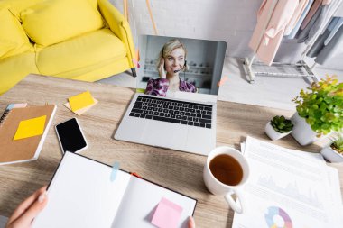Kulaklıkla gülümseyen kadın, çay fincanının yanında dizüstü bilgisayar ekranında ve Webinar sırasında elinde not defteri olan serbest çalışan bulanık ön planda. 