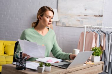 Blonde Freelancer elinde belgelerle bardağın yanında dizüstü bilgisayar ve bulanık ön planda bitkiler kullanıyor. 