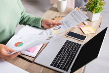 Cropped view of teleworker holding documents with charts near devices with blank screen on blurred foreground  clipart