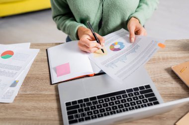 Cropped view of teleworker writing on sticky note on document with charts near notebook and laptop on blurred foreground  clipart