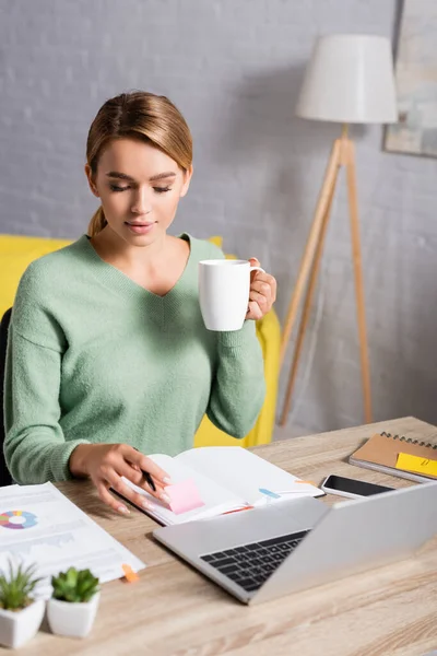 Freelancer Sosteniendo Taza Mientras Mira Cuaderno Cerca Dispositivos Papeles Primer — Foto de Stock