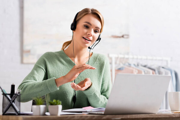 Smiling freelancer in headset gesturing during video chat on laptop on blurred foreground at home 