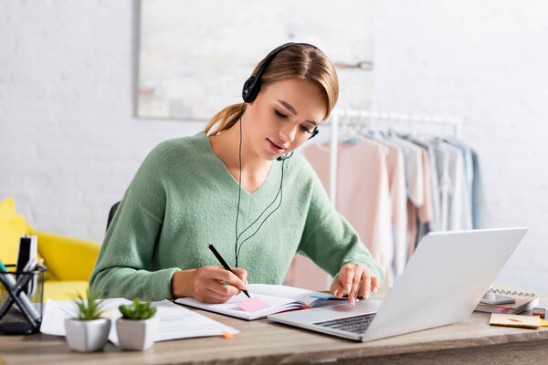 Freelancer in headset using laptop and writing on notebook during video call at home 